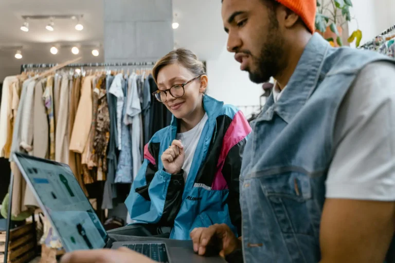 Homens e mulher sorrindo enquanto olham para um laptop em uma loja de roupas, explorando opções de compra, com prateleiras de roupas ao fundo.